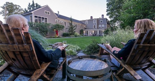 Two people relax in wooden chairs, holding drinks, overlooking a lush garden and a large house in the background.