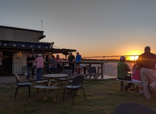 A sunset view over water with people socializing at an outdoor bar, surrounded by tables and chairs.