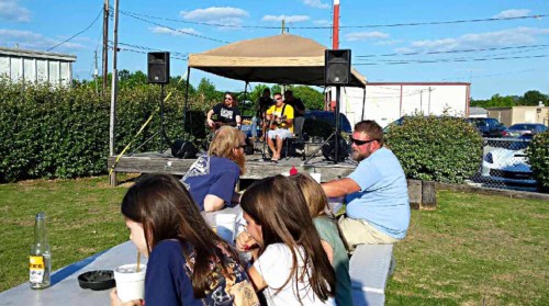 A band performs on stage while people sit at a table enjoying food outdoors on a sunny day.
