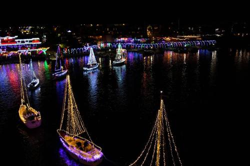 Colorful boats adorned with lights float on a dark waterway, surrounded by festive decorations along the shore.