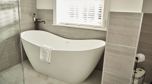 A modern bathroom featuring a sleek, freestanding white bathtub and gray tiled walls. Natural light filters through a window.