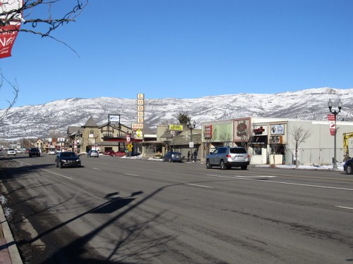A snowy mountain backdrop frames a busy street lined with shops and cars under a clear blue sky.