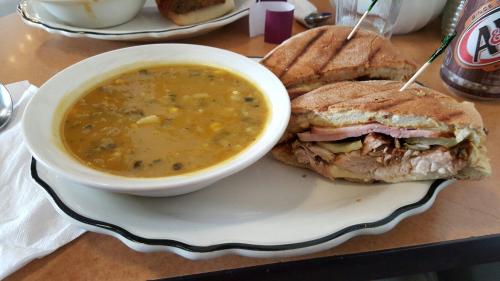 A plate with a bowl of yellow soup and a sandwich filled with meat and pickles, garnished with toothpicks.