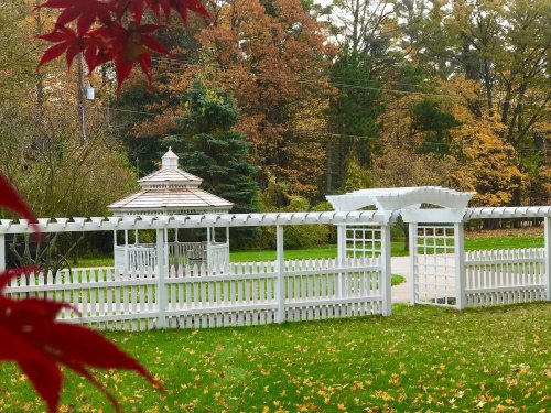 A white gazebo surrounded by a picket fence, set in a colorful autumn landscape with trees and fallen leaves.