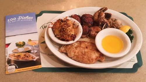 A plate of seafood and fried fish with a side of dipping sauce, accompanied by a brochure about Alabama dishes.