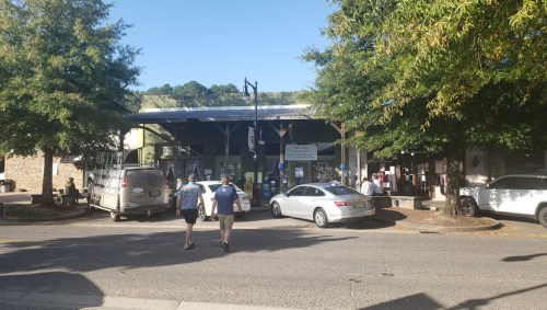 Two people walk towards a market area with trees and parked cars under a clear blue sky.