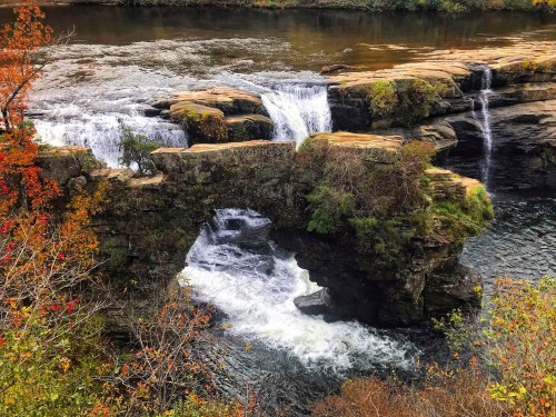 A scenic view of a stone arch bridge over a river with waterfalls, surrounded by autumn foliage.