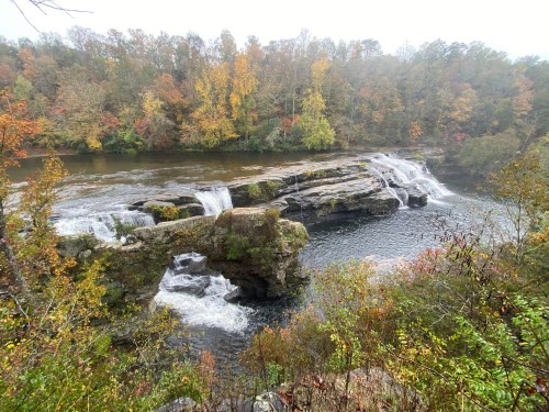 A scenic view of a waterfall cascading over rocks, surrounded by colorful autumn foliage and a calm river.