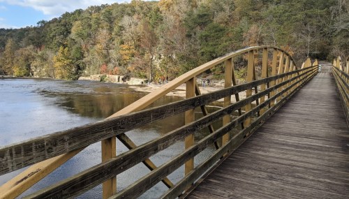 A wooden bridge arches over a calm river, surrounded by trees with autumn foliage.