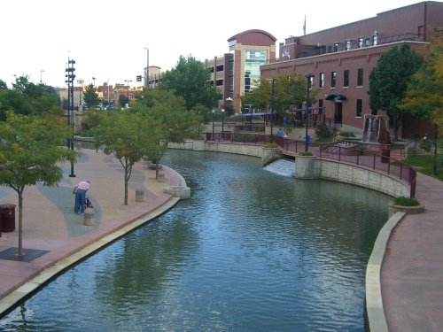 A serene park with a winding waterway, trees lining the path, and buildings in the background. People stroll along the walkway.