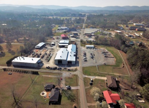 Aerial view of a rural area featuring buildings, vehicles, and trees, with mountains in the background.