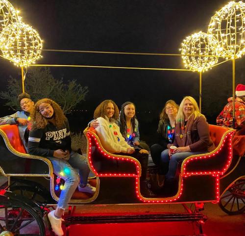 A group of six women sitting in a festive, illuminated sleigh decorated with lights, smiling at night.