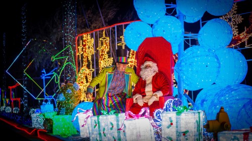 A colorful holiday scene featuring a man in a top hat sitting next to Santa Claus amidst bright lights and decorated gifts.