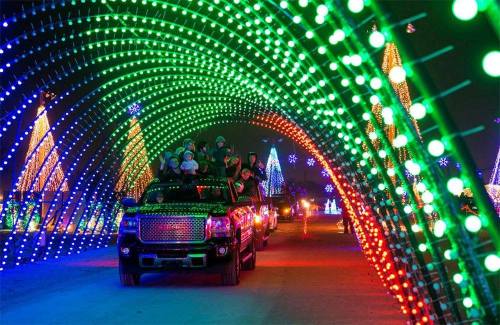 A festive scene with a truck driving through a colorful light tunnel, surrounded by illuminated trees and decorations.