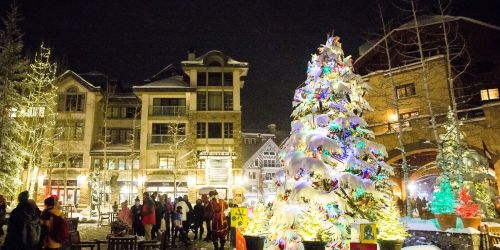 A festive scene with a brightly lit Christmas tree and people walking in a charming, snow-covered village at night.