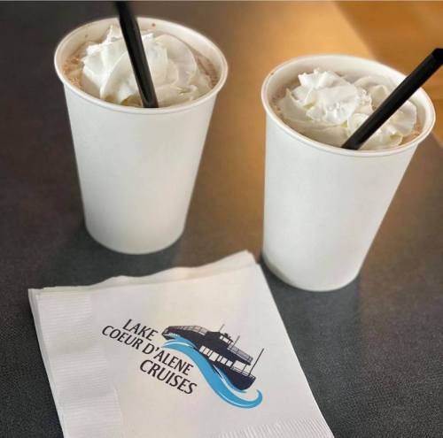 Two cups of whipped cream-topped drinks with black straws, next to a napkin featuring "Lake Coeur d'Alene Cruises."