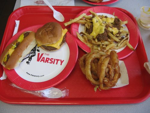 A red tray with a cheeseburger, hot dog, loaded fries, and onion rings on paper plates.