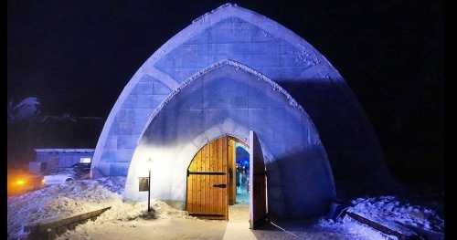 A large, illuminated ice structure with a wooden door, surrounded by snow at night.