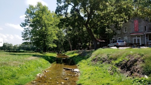 A serene landscape featuring a clear stream, lush greenery, and a rustic building in the background under a blue sky.