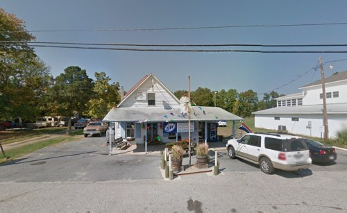 A small, two-story building with a porch, surrounded by trees and parked cars, featuring a sign for Miller Lite.