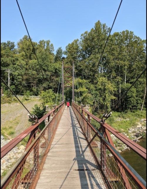 A person walks across a suspension bridge surrounded by trees and a river under a clear blue sky.