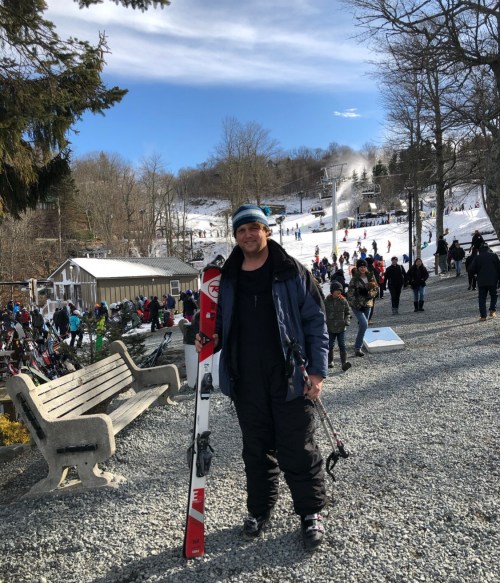 A person stands on a gravel path holding skis, with a ski resort and snow-covered slopes in the background.
