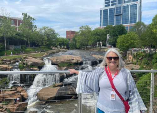 A woman with long gray hair stands by a waterfall, smiling, with a city skyline in the background.