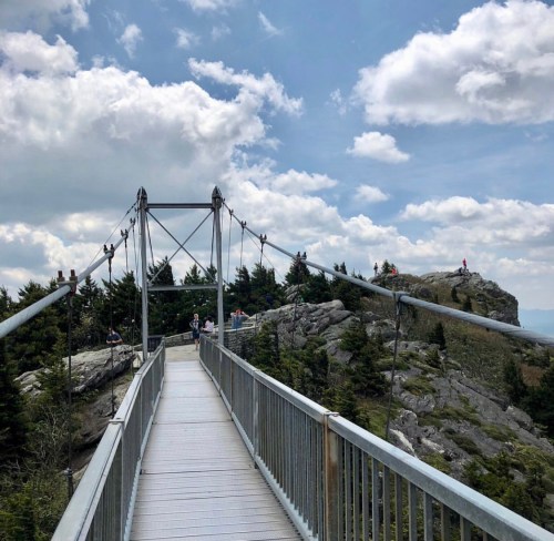 A suspension bridge leads to a rocky outcrop surrounded by trees, with people enjoying the view under a cloudy sky.