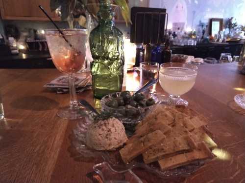 A cozy table setting with drinks, olives, and a plate of crackers and cheese, illuminated by soft candlelight.