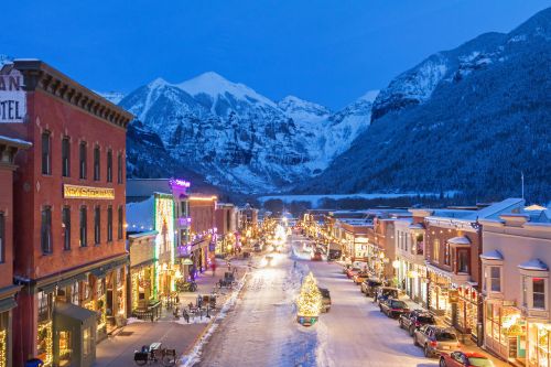 A snowy mountain town illuminated with festive lights, featuring shops and a decorated Christmas tree under a twilight sky.