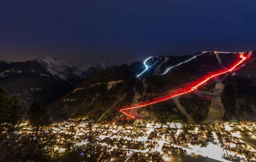 A nighttime view of a snowy mountain with ski trails lit up in red and blue, overlooking a brightly lit town below.
