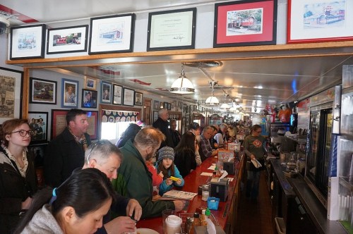 A busy diner with a long counter, patrons enjoying meals, and walls adorned with framed photos and certificates.