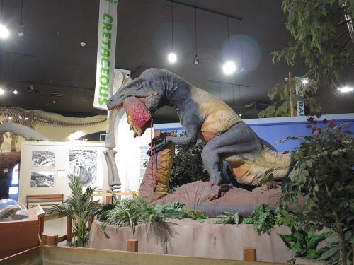 A large dinosaur model stands in a museum exhibit, surrounded by plants and educational displays about prehistoric life.