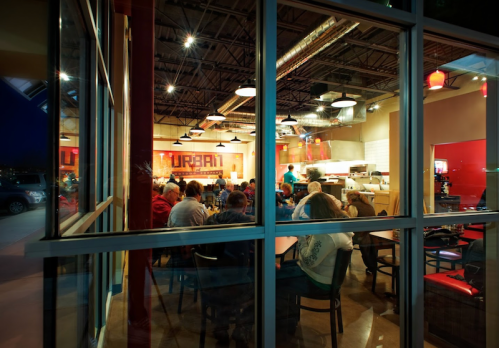 A bustling restaurant interior with diners gathered around tables, seen through large windows at night.