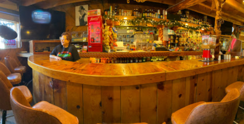A cozy bar with wooden decor, featuring a bartender and shelves stocked with various drinks.