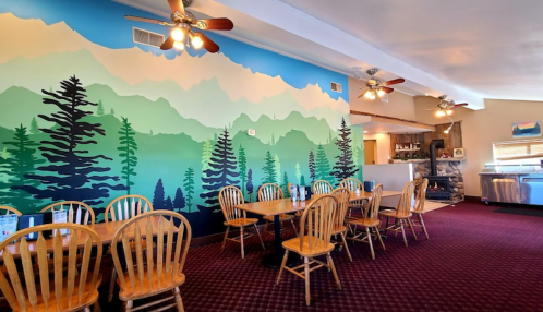 A cozy restaurant interior featuring wooden tables, chairs, and a colorful mountain mural on the wall.