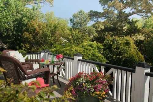 A cozy balcony with wicker furniture, flowers, and a view of lush greenery and trees in the background.