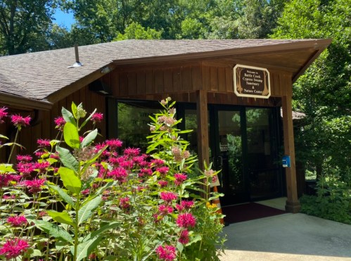 A wooden nature center building surrounded by vibrant pink flowers and lush greenery on a sunny day.