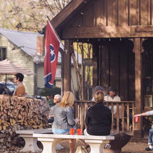 A group of people sitting at a table outside a rustic building, with a Tennessee flag and stacked firewood nearby.