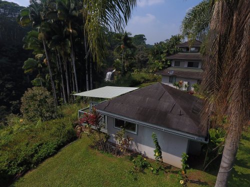 A serene landscape featuring a house surrounded by lush greenery and palm trees, with a waterfall in the background.