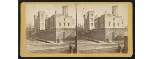 Historic black-and-white photo of a large building with towers, set on a cobblestone street, featuring shadows and a figure.