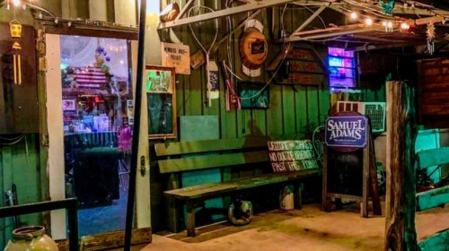 A rustic bar entrance with colorful lights, wooden benches, and various signs, creating a lively atmosphere.