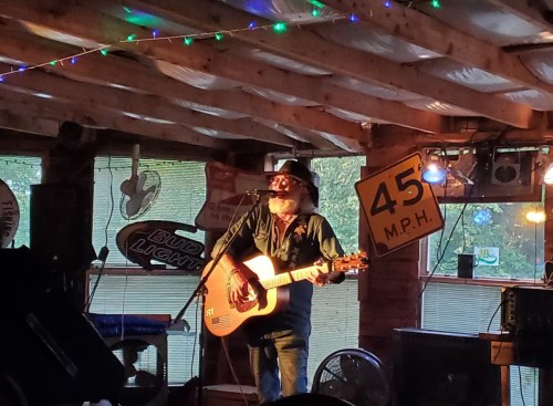A musician with a beard and hat performs on stage with an acoustic guitar in a cozy, decorated venue.