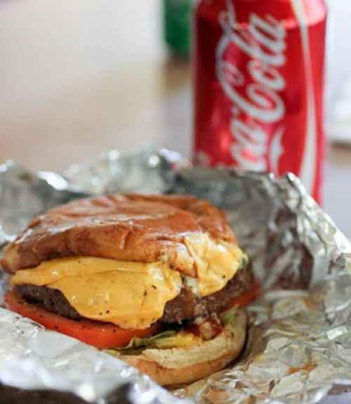 A cheeseburger with lettuce and tomato, wrapped in foil, next to a can of Coca-Cola.