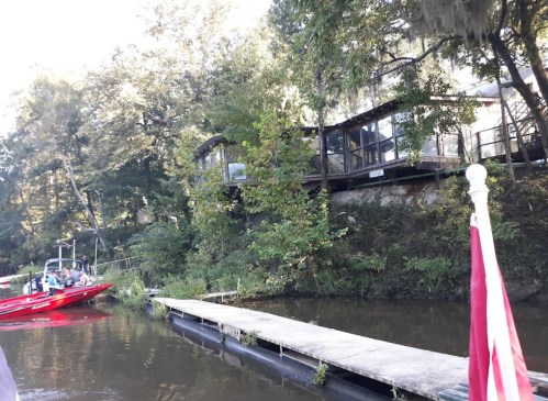 A view of a riverbank with a wooden dock, a boat, and a house surrounded by trees.
