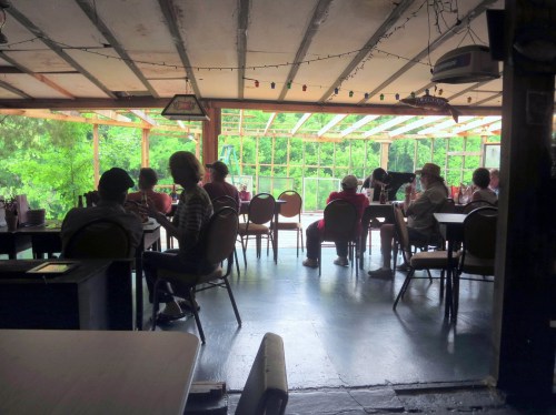 A rustic dining area with people seated at tables, large windows showing greenery outside.