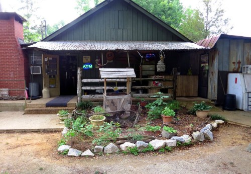 A rustic building with a metal roof, surrounded by a small garden and potted plants, featuring an ATM sign.