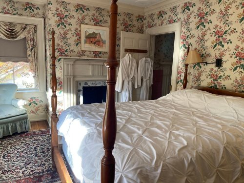 Cozy bedroom with floral wallpaper, a four-poster bed, and two white robes hanging by the fireplace.