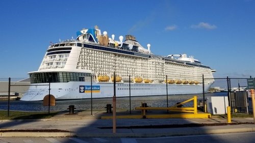 A large cruise ship docked at a port, featuring multiple decks and yellow lifeboats, under a clear blue sky.