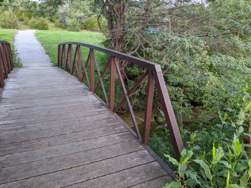 A wooden bridge with a metal railing spans a small creek, surrounded by lush greenery and a pathway.